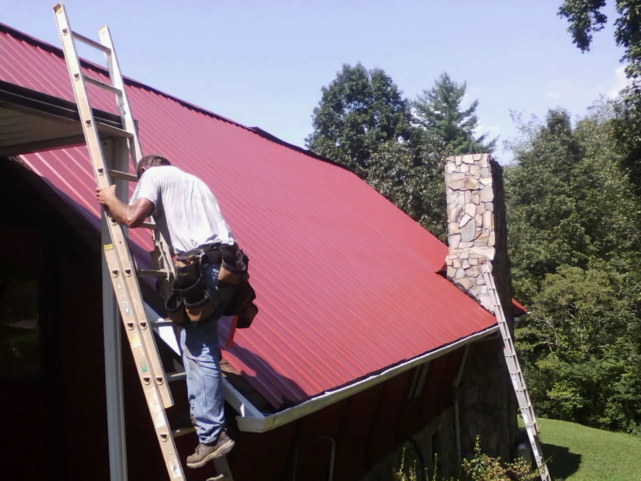 Roofer on a ladder beside a red metal roof and stone chimney — Soffit & Fascia Repair by Roofing Brothers in Maugansville